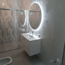 Piter HOME - Nowoczesna łazienka z owalnym lustrem LED, białą szafką pod umywalką i kabiną prysznicową z wzorzystymi płytkami. Minimalistyczny design, szare płytki podłogowe.