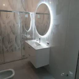 Nowoczesna łazienka z owalnym lustrem LED, białą szafką pod umywalką i kabiną prysznicową z wzorzystymi płytkami. Minimalistyczny design, szare płytki podłogowe.