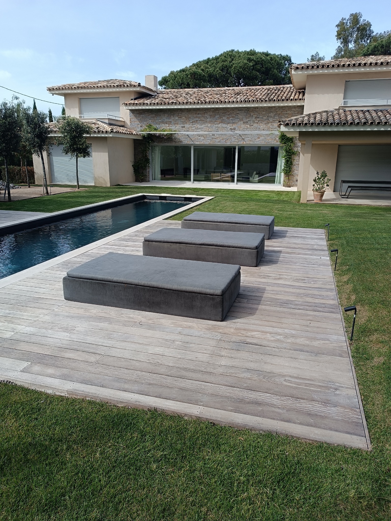 Nowoczesny dom z tarasem z drewna, basenem i zadbanym trawnikiem w słoneczny dzień. Widoczne trzy szare leżanki na tarasie. Minimalistyczny design.