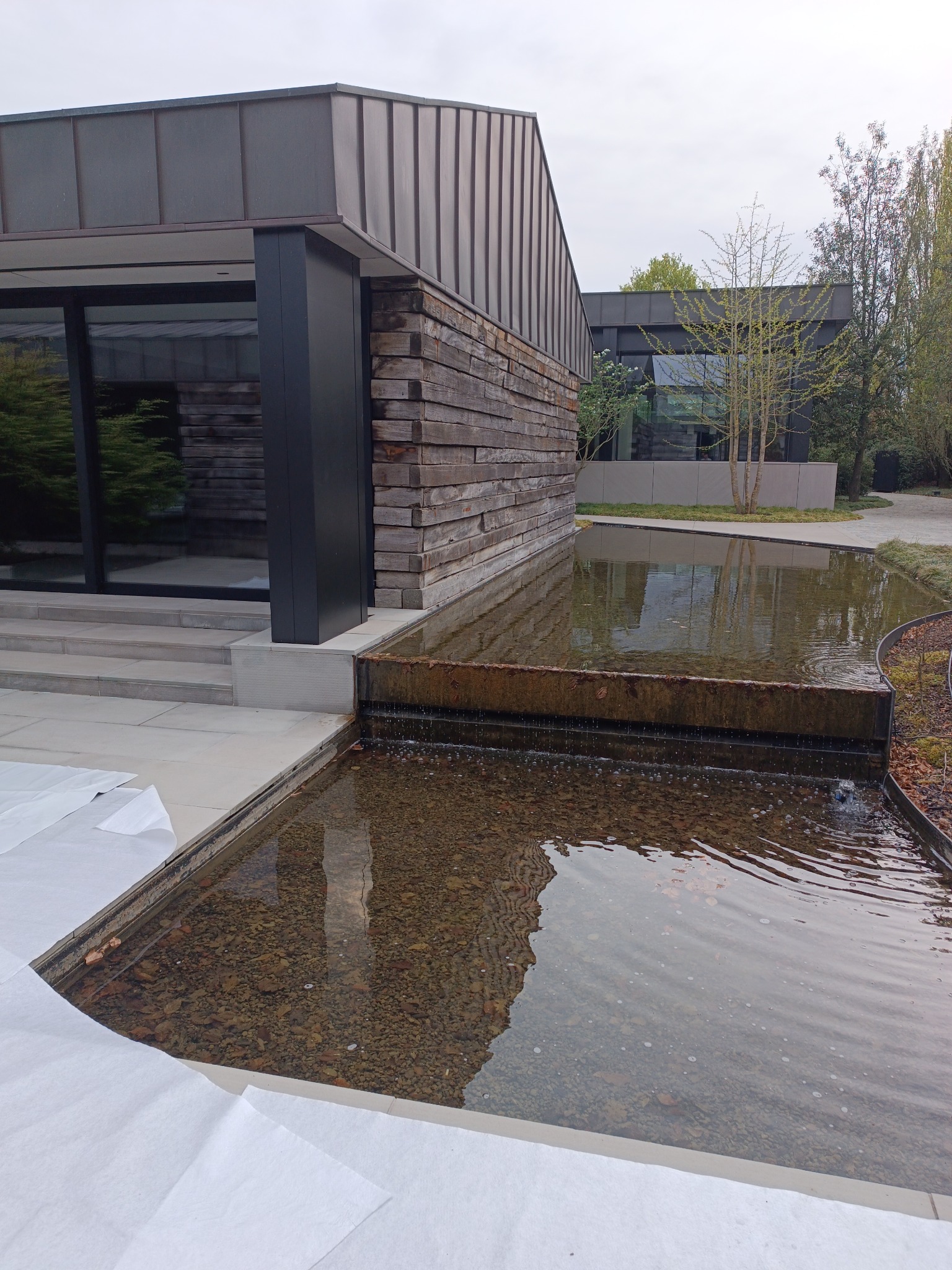 Nowoczesny budynek z drewnianą elewacją i metalowym dachem, obok niego zbiornik wodny z kaskadą. Część powierzchni zabezpieczona białą folią.