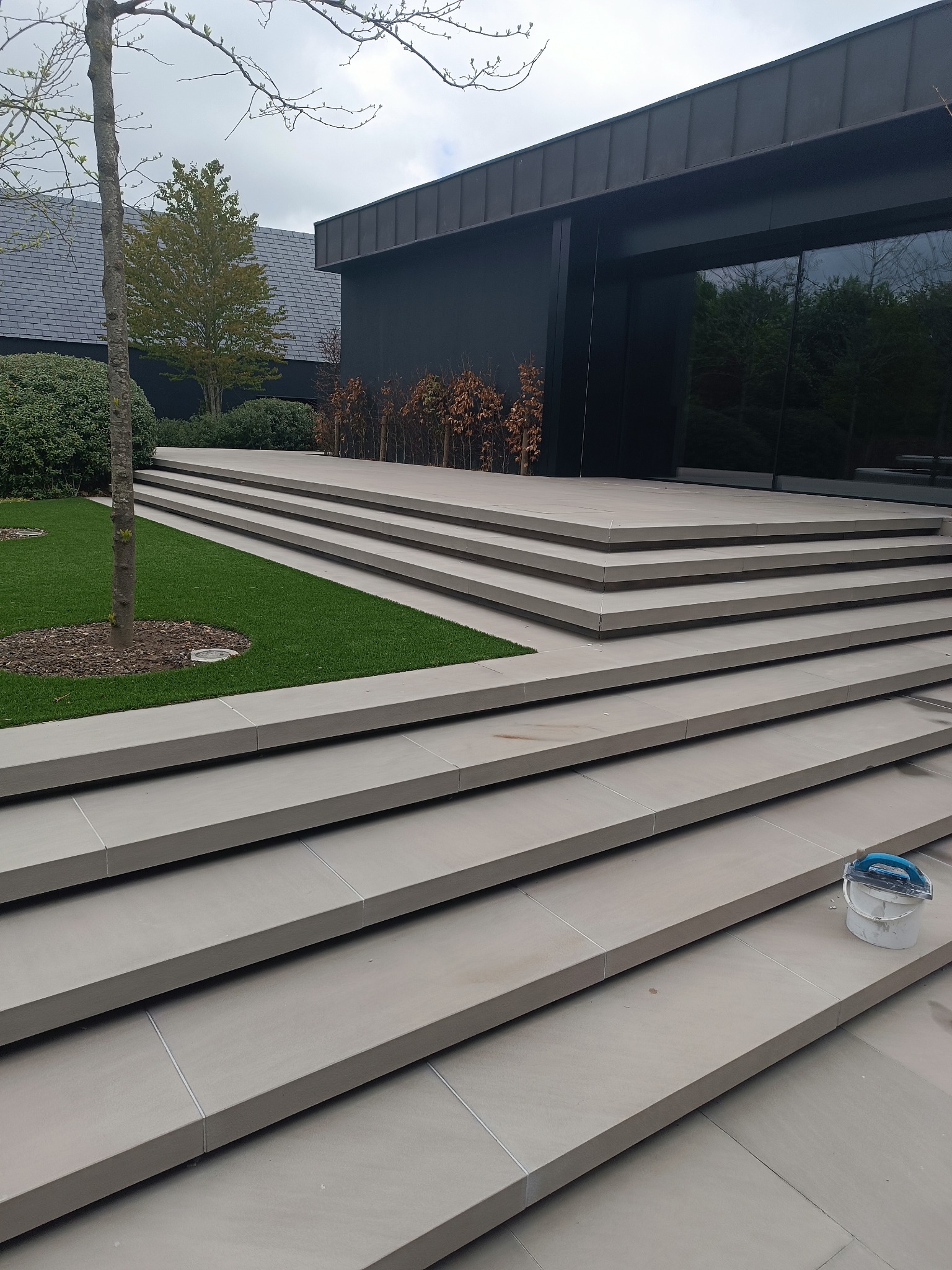 Eleganckie, zewnętrzne schody z jasnych, prostokątnych płyt prowadzące do nowoczesnego budynku z ciemną elewacją i dużymi oknami. Widoczny fragment trawnika i drzewo.
