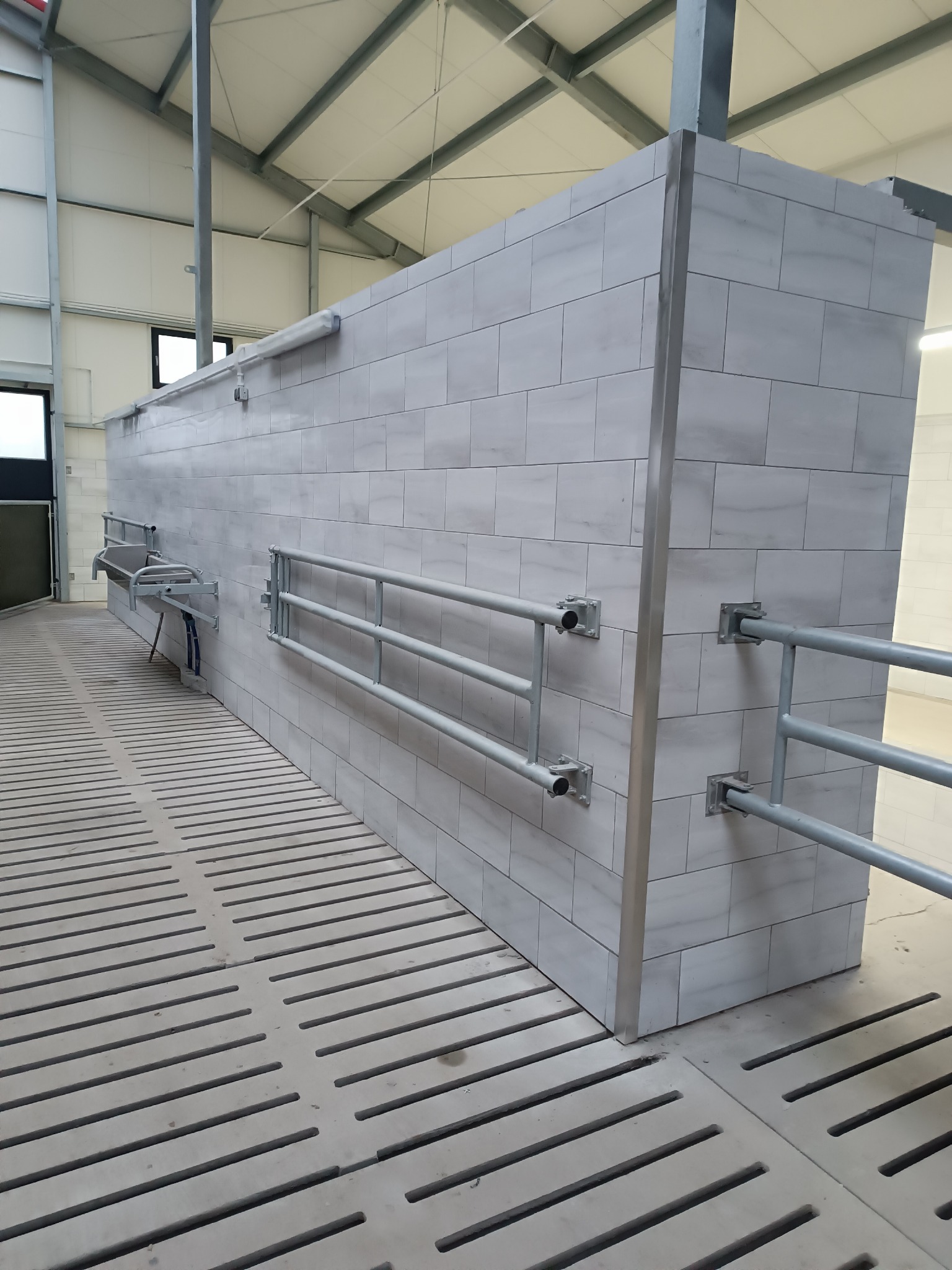 Wykończona ściana w hali z jasnych płytek imitujących marmur, z zamontowanymi metalowymi barierkami i umywalką. Podłoga z ażurowych płyt betonowych.