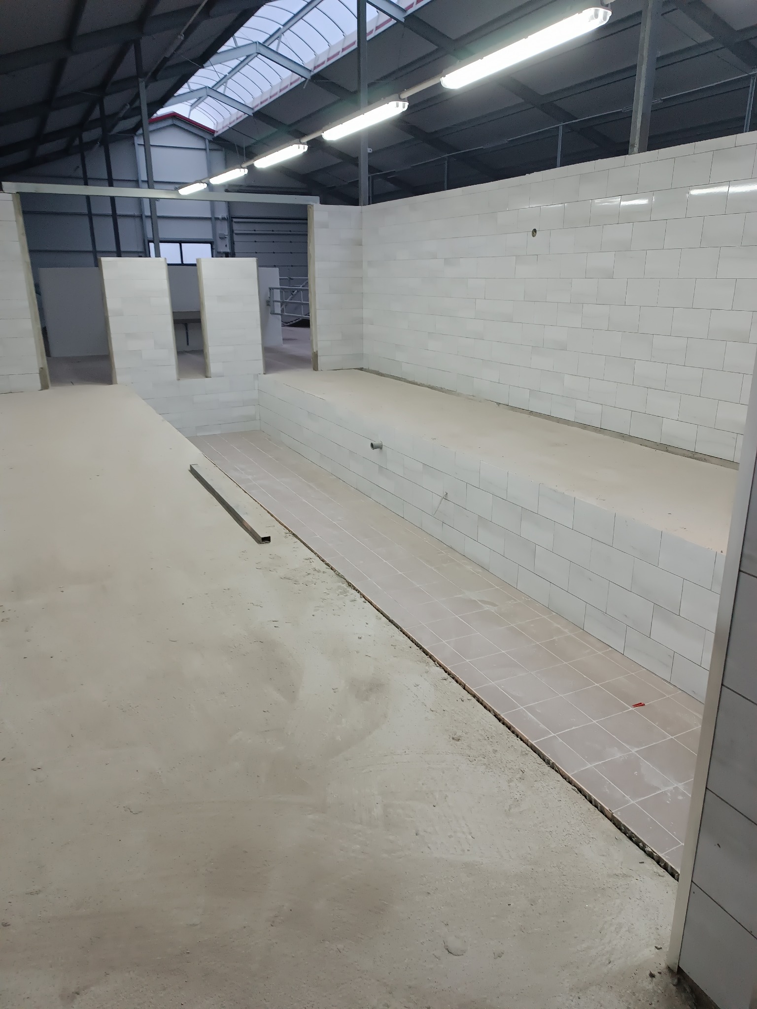 Wnętrze w trakcie prac wykończeniowych: częściowo wyłożone płytki na ścianach i podłodze, surowy beton na posadzce, widoczne przygotowania do dalszych prac.