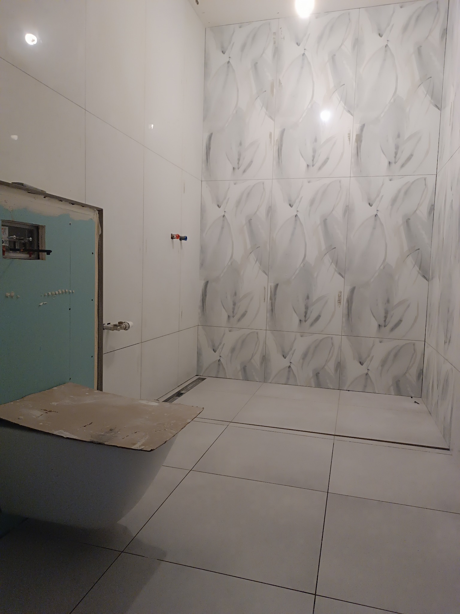 Łazienka w trakcie wykańczania: białe płytki z motywem liści na ścianach, częściowo zabudowany stelaż WC. Widoczny odpływ liniowy w podłodze.