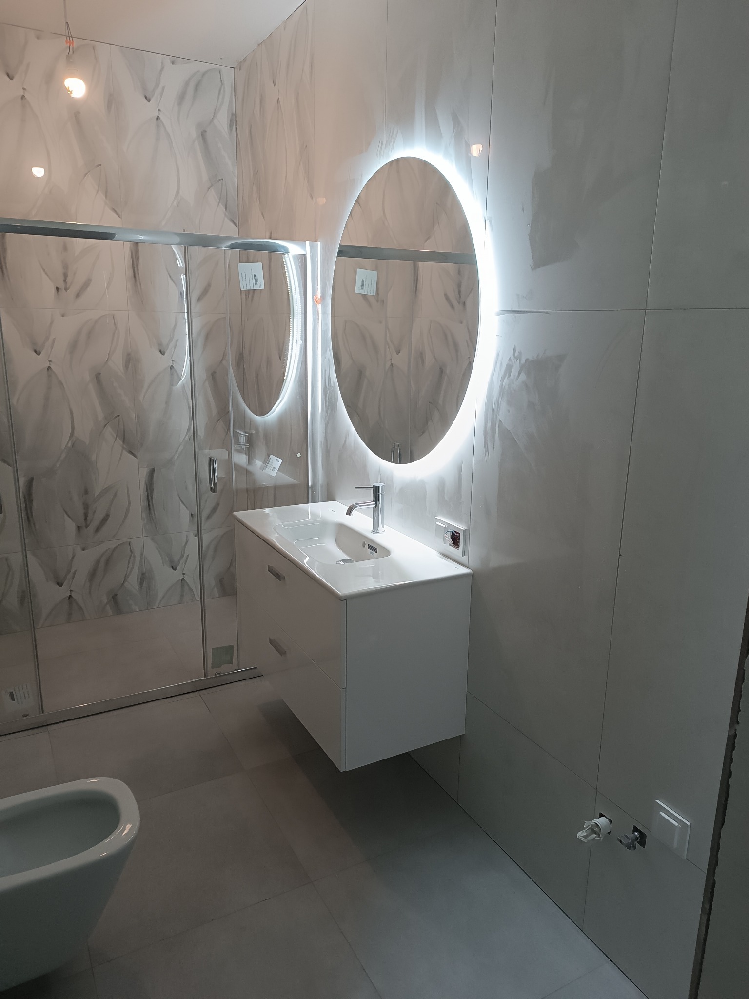 Nowoczesna łazienka z owalnym lustrem LED, białą szafką pod umywalką i kabiną prysznicową z wzorzystymi płytkami. Minimalistyczny design, szare płytki podłogowe.