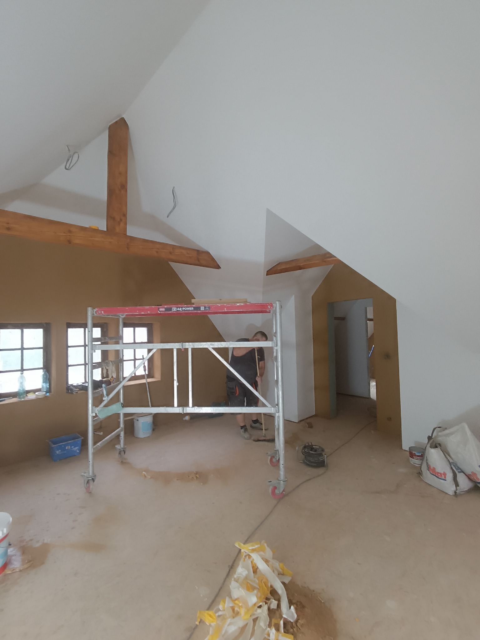 Poddasze w trakcie prac. Widoczny fragment rusztowania, otynkowana ściana w kolorze piaskowym oraz pomalowany na biało sufit ze skośnymi belkami.