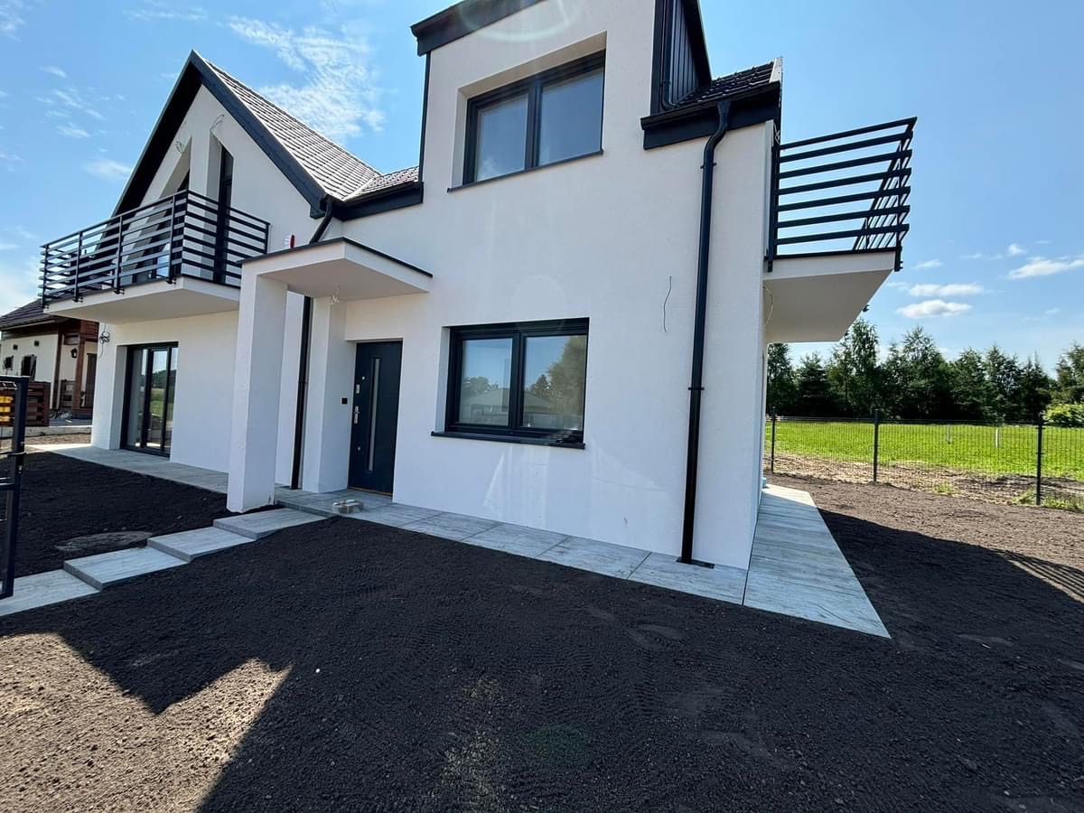 Nowoczesny, dwukondygnacyjny dom z białymi ścianami, czarnymi ramami okiennymi i balkonami z czarnymi balustradami. Teren przed domem przygotowany pod trawnik, z ułożonymi płytami chodnikowymi...