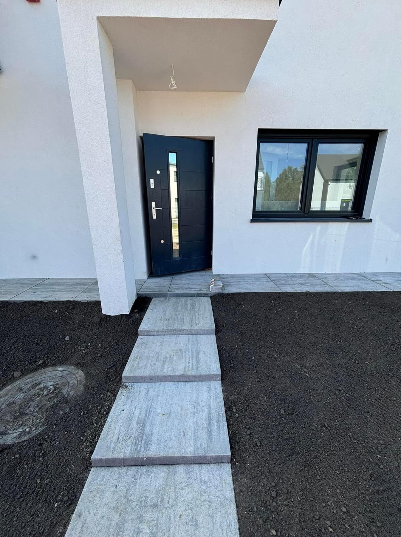 Nowoczesny dom z otwartymi, grafitowymi drzwiami wejściowymi i czarną ramą okienną, z betonowymi płytami tworzącymi ścieżkę przez świeżo rozplantowaną ziemię.