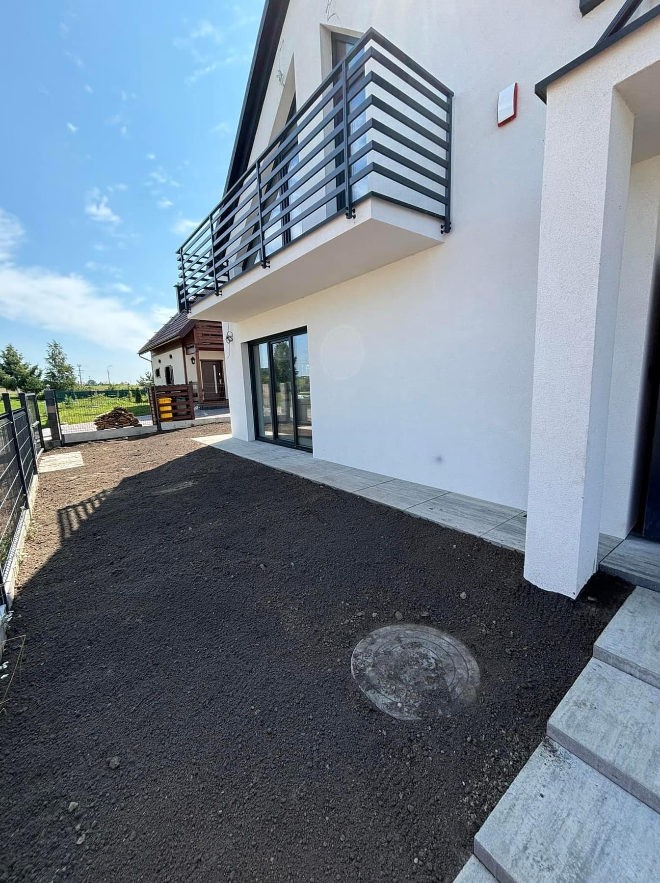 Przygotowany teren pod trawnik przy nowoczesnym domu z balkonem i czarną stolarką okienną, widoczna studzienka kanalizacyjna