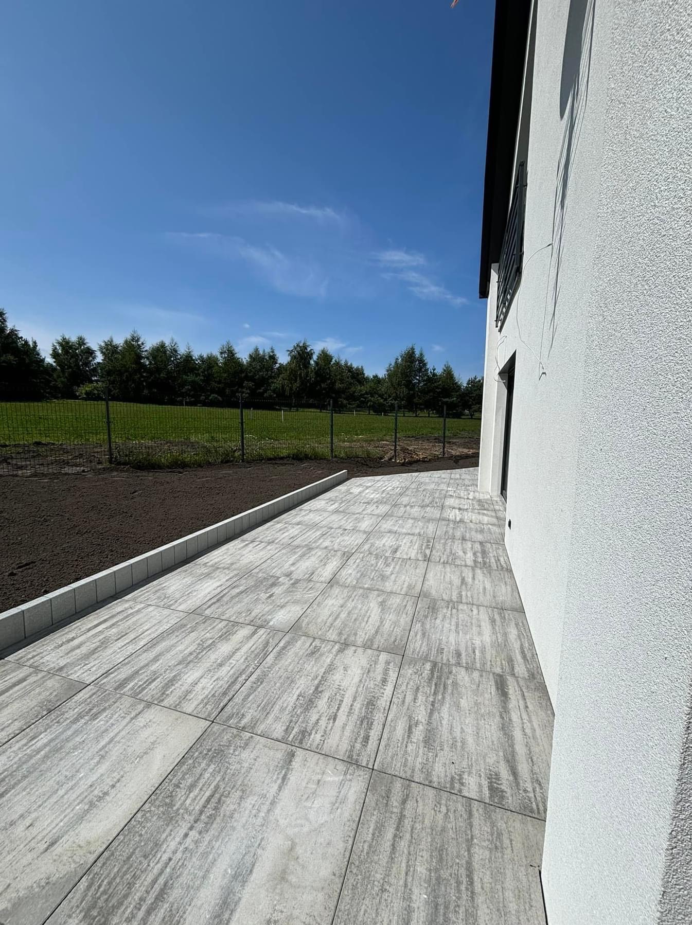 Ułożona szara kostka brukowa o wzorze imitującym drewno, przylegająca do białego budynku z widoczną siatką ogrodzeniową i przygotowanym terenem pod trawnik w tle.