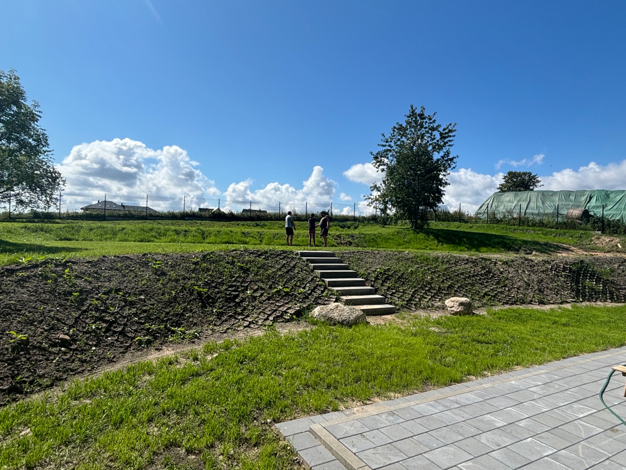 Ujęcie krajobrazu z betonowymi schodami prowadzącymi na wzgórze, na którym stoją trzy osoby, z widocznym systemem zabezpieczenia skarpy, trawnikiem i fragmentem utwardzonej powierzchni z kostki...