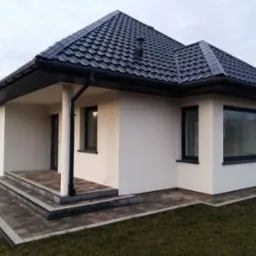 Nowoczesny parterowy dom z ciemną dachówką, białymi ścianami i kostką brukową przed wejściem, sfotografowany w pochmurny dzień.