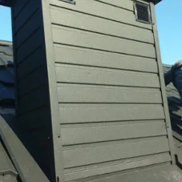 Szczegółowe ujęcie komina wentylacyjnego z metalową obudową w kolorze grafitowym, z widocznymi otworami wentylacyjnymi i fragmentem dachu pokrytego blachodachówką, na tle błękitnego nieba.