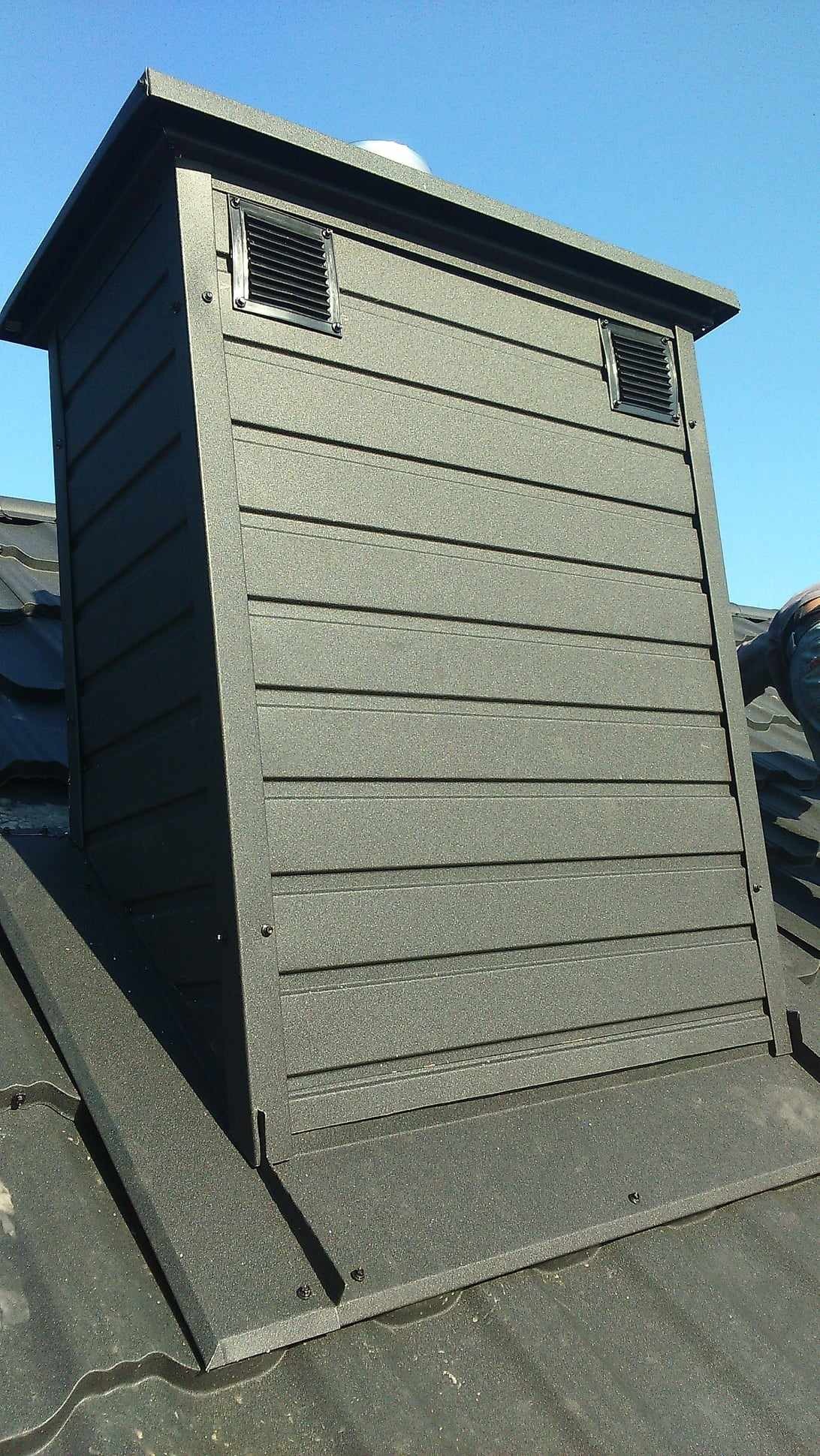 Szczegółowe ujęcie komina wentylacyjnego z metalową obudową w kolorze grafitowym, z widocznymi otworami wentylacyjnymi i fragmentem dachu pokrytego blachodachówką, na tle błękitnego nieba.