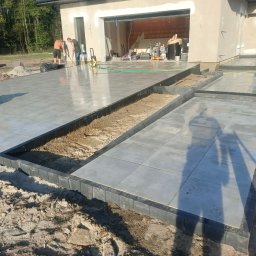 Dobra Ekipa - Ułożona, szara, błyszcząca kostka na tarasie domu w trakcie prac wykończeniowych. Na pierwszym planie czarne obrzeża. Dwóch mężczyzn czyści powierzchnię.