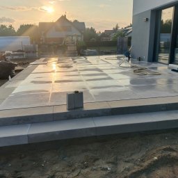 Dobra Ekipa - Jasnoszara, duża kostka na tarasie domu jednorodzinnego w słoneczny dzień. Widoczny fragment elewacji, traktor i bus z kostką w tle.