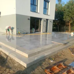 Czyszczenie tarasu z płyt gresowych po montażu, z widocznymi schodami i fragmentem domu w tle. Prace wykończeniowe na zewnątrz budynku.