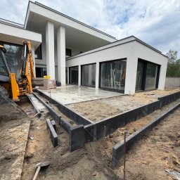 Dobra Ekipa - Przygotowanie podłoża pod taras lub ścieżki przy nowoczesnym białym domu, z koparką JCB, czarnymi obrzeżami i piaskową warstwą wyrównującą.