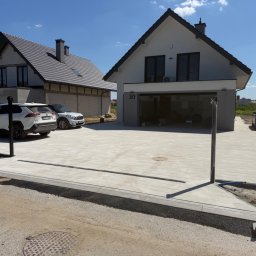 Dobra Ekipa - Jasnoszary podjazd z prostokątnych płyt betonowych przed nowoczesnym domem z garażem, z widocznym liniowym odpływem. Obok dom w budowie.
