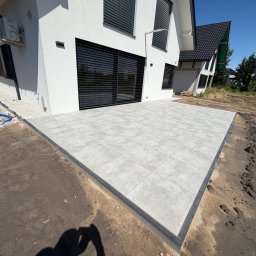 Dobra Ekipa - Nowoczesny taras z dużych, jasnoszarych płyt betonowych z ciemnym obrzeżem, przylegający do białego domu z czarnymi żaluzjami, na tle niezagospodarowanego gruntu.