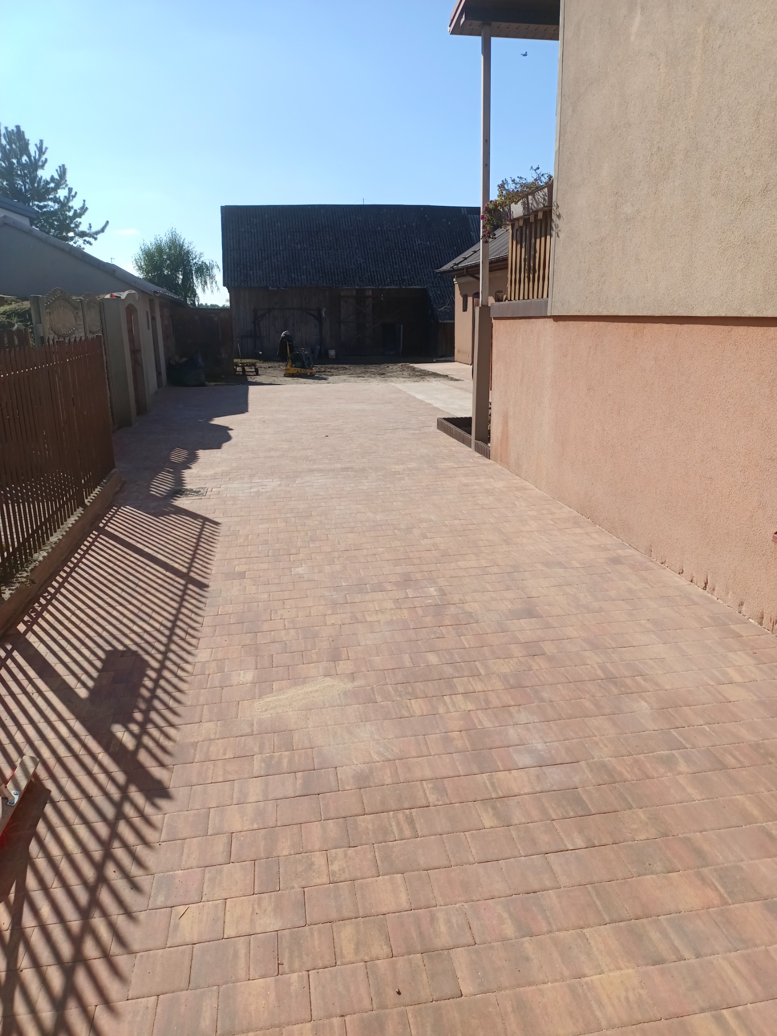 Ułożona, brązowa kostka brukowa na podjeździe, widok z perspektywy. W tle budynek gospodarczy i sprzęt budowlany. Cień ogrodzenia na kostce. Słoneczny dzień.