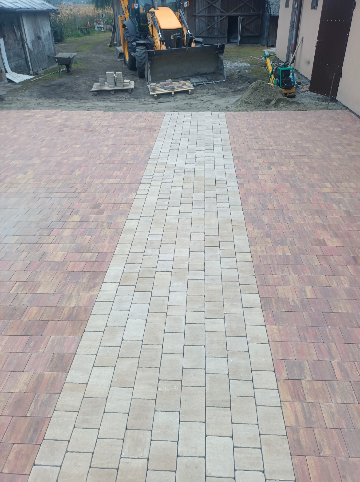 Ukończona nawierzchnia z kostki brukowej w odcieniach beżu i brązu, z widocznym traktorem i zagęszczarką w tle. Estetyczne wykończenie podjazdu.