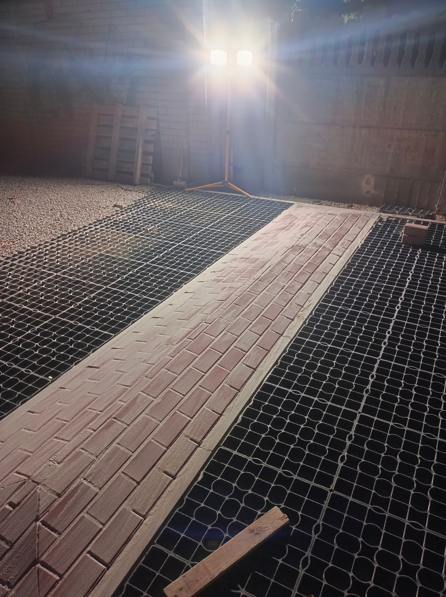 Częściowo ułożona ścieżka z czerwonej kostki brukowej na czarnej siatce, oświetlona lampą budowlaną. Widoczne narzędzia i materiały budowlane.