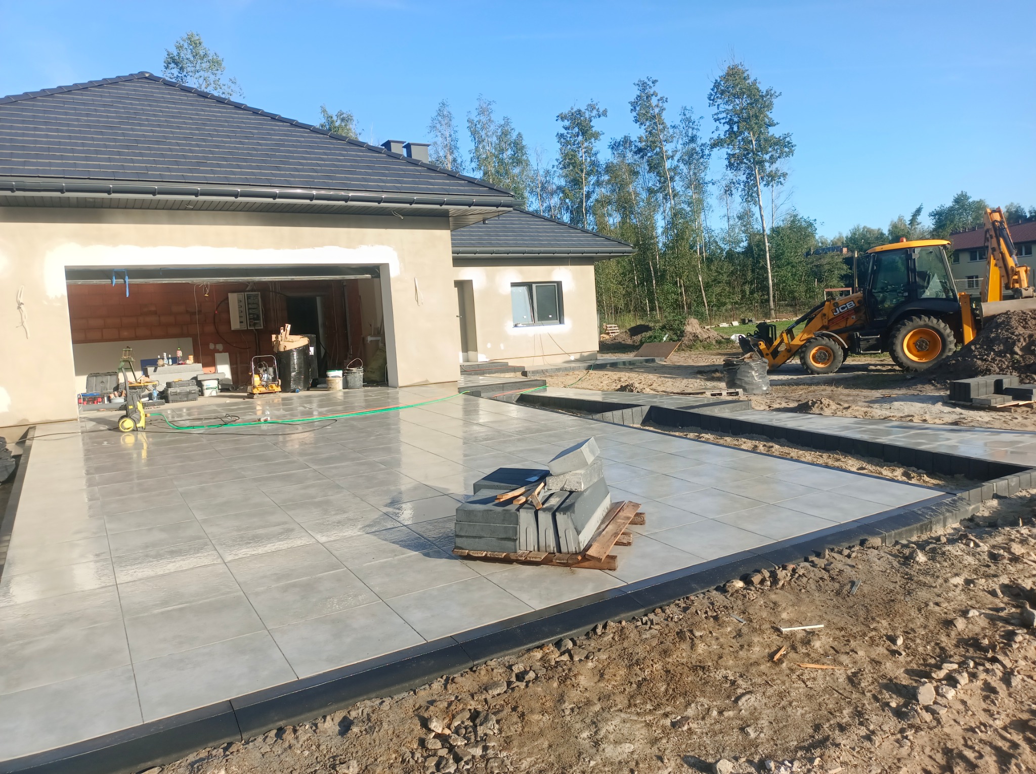 Układanie jasnoszarej kostki brukowej na posesji domu jednorodzinnego. W tle widoczny żółty traktor JCB i drzewa.