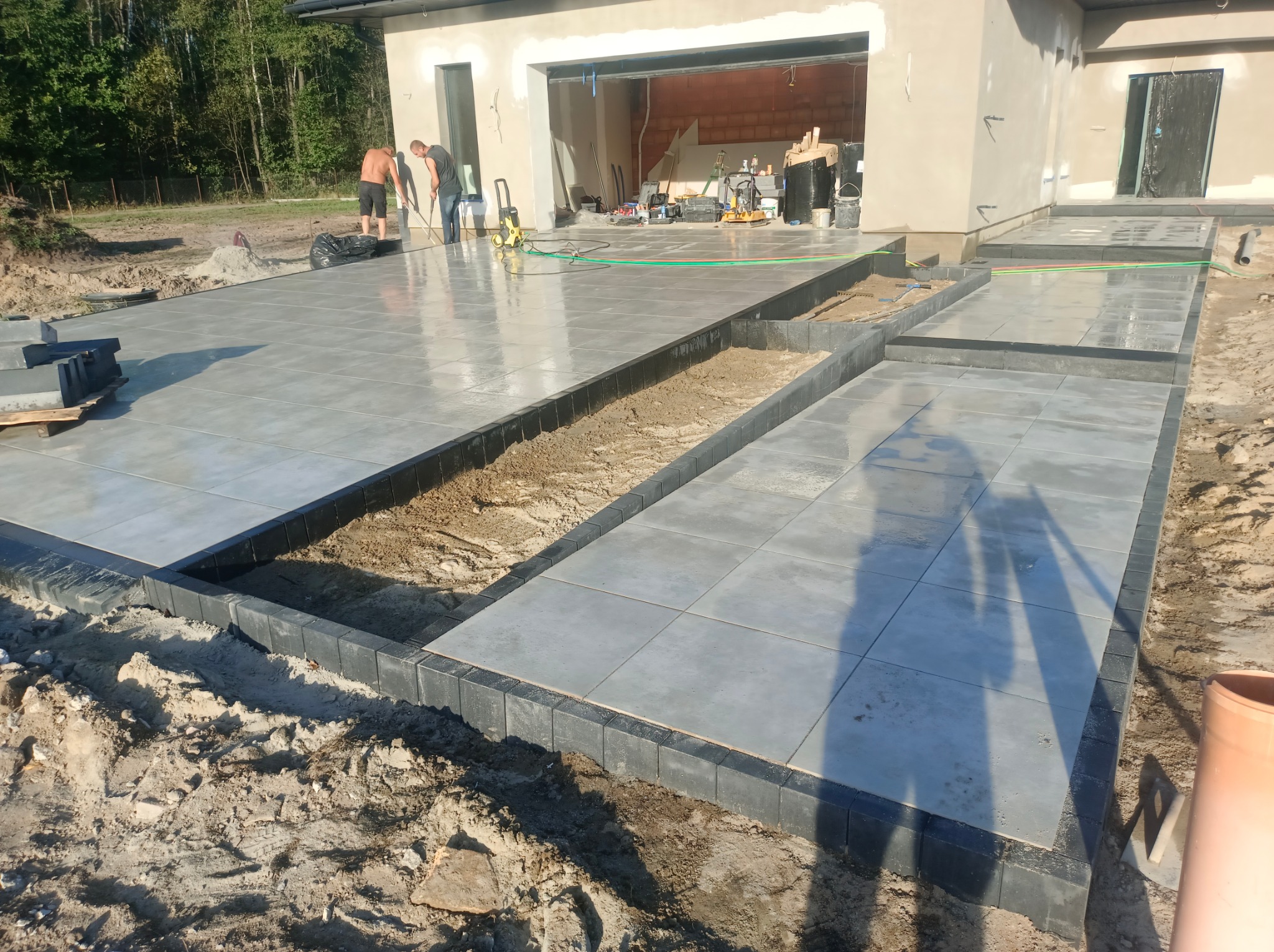 Ułożona, szara, błyszcząca kostka na tarasie domu w trakcie prac wykończeniowych. Na pierwszym planie czarne obrzeża. Dwóch mężczyzn czyści powierzchnię.