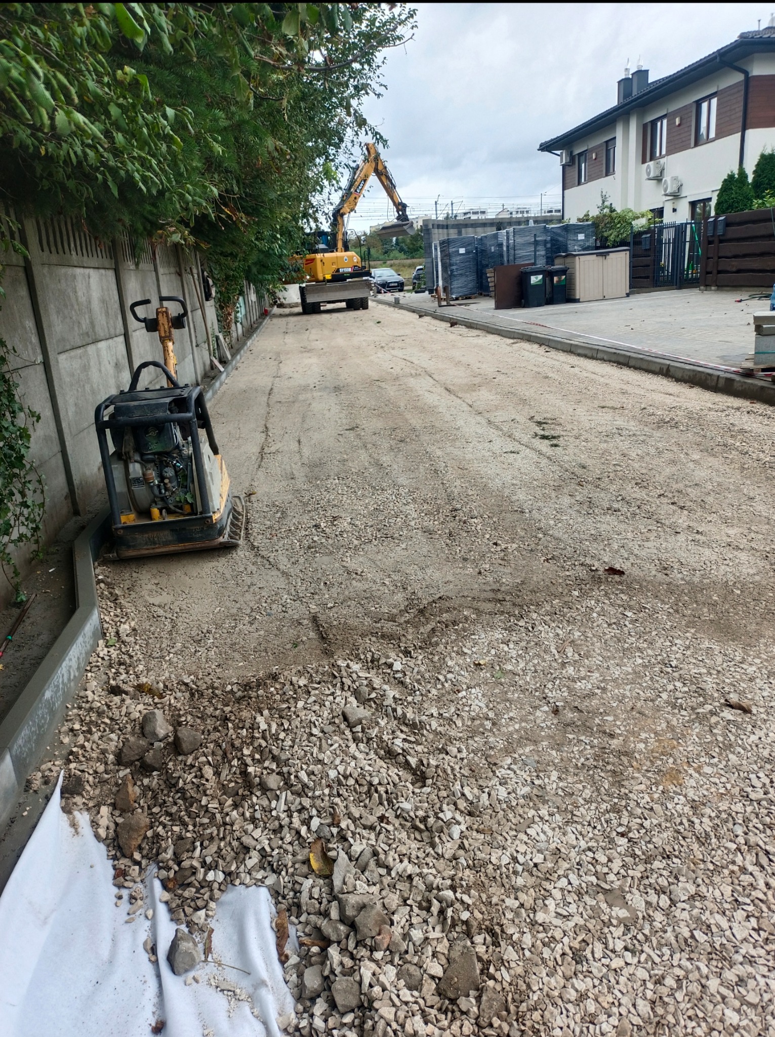 Przygotowanie podłoża: ubijarka wibrująca obok krawężnika, w tle koparka i stosy kostki, dom jednorodzinny, pochmurne niebo.