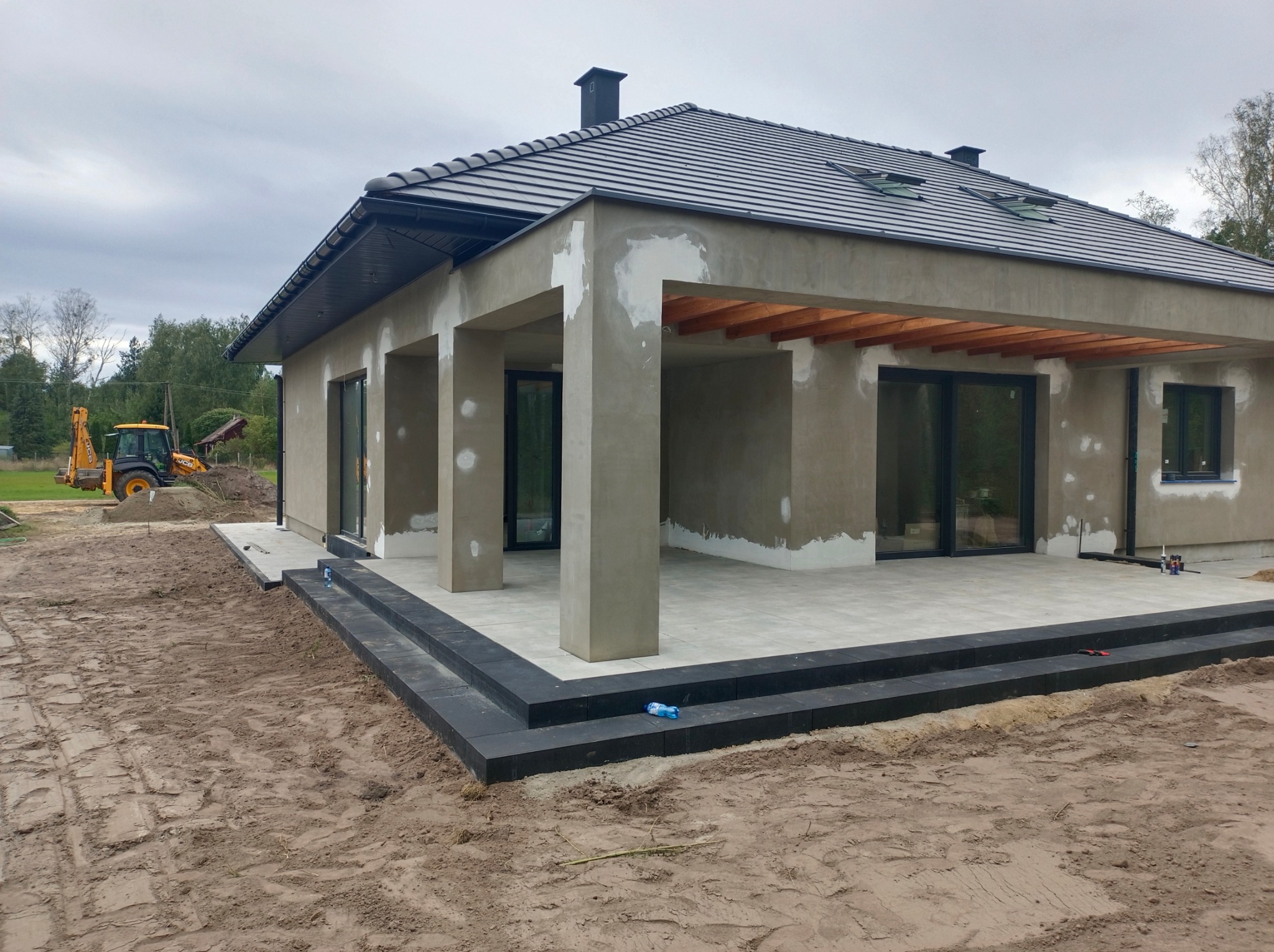 Nowoczesny taras z jasnej kostki i czarnych obrzeży przed domem w trakcie budowy. W tle widoczny sprzęt budowlany i częściowo otynkowana elewacja.