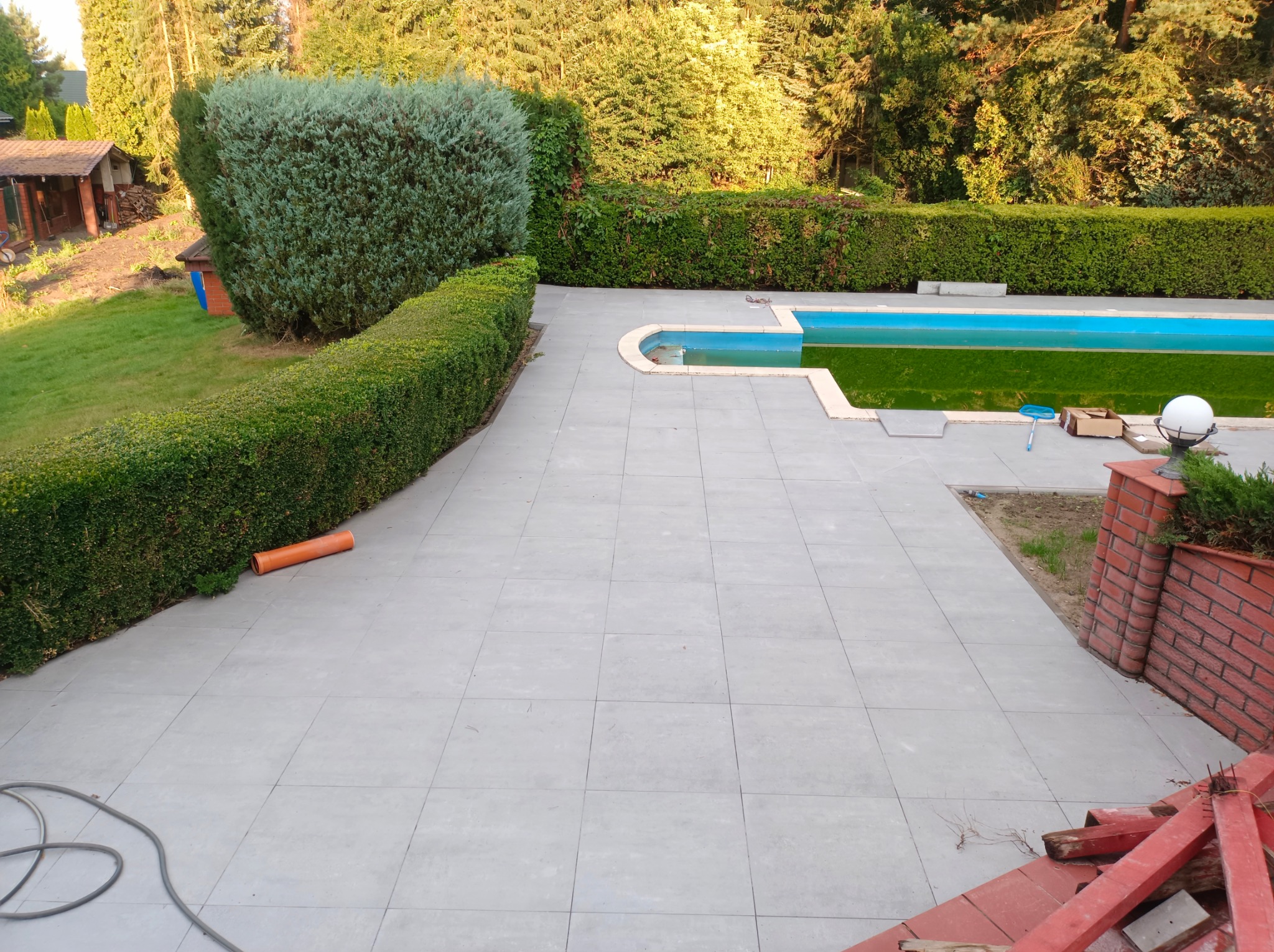 Jasnoszara kostka brukowa wokół basenu, otoczona zielenią i żywopłotem. Fragment czerwonej cegły i elementy budowlane w prawym dolnym rogu.