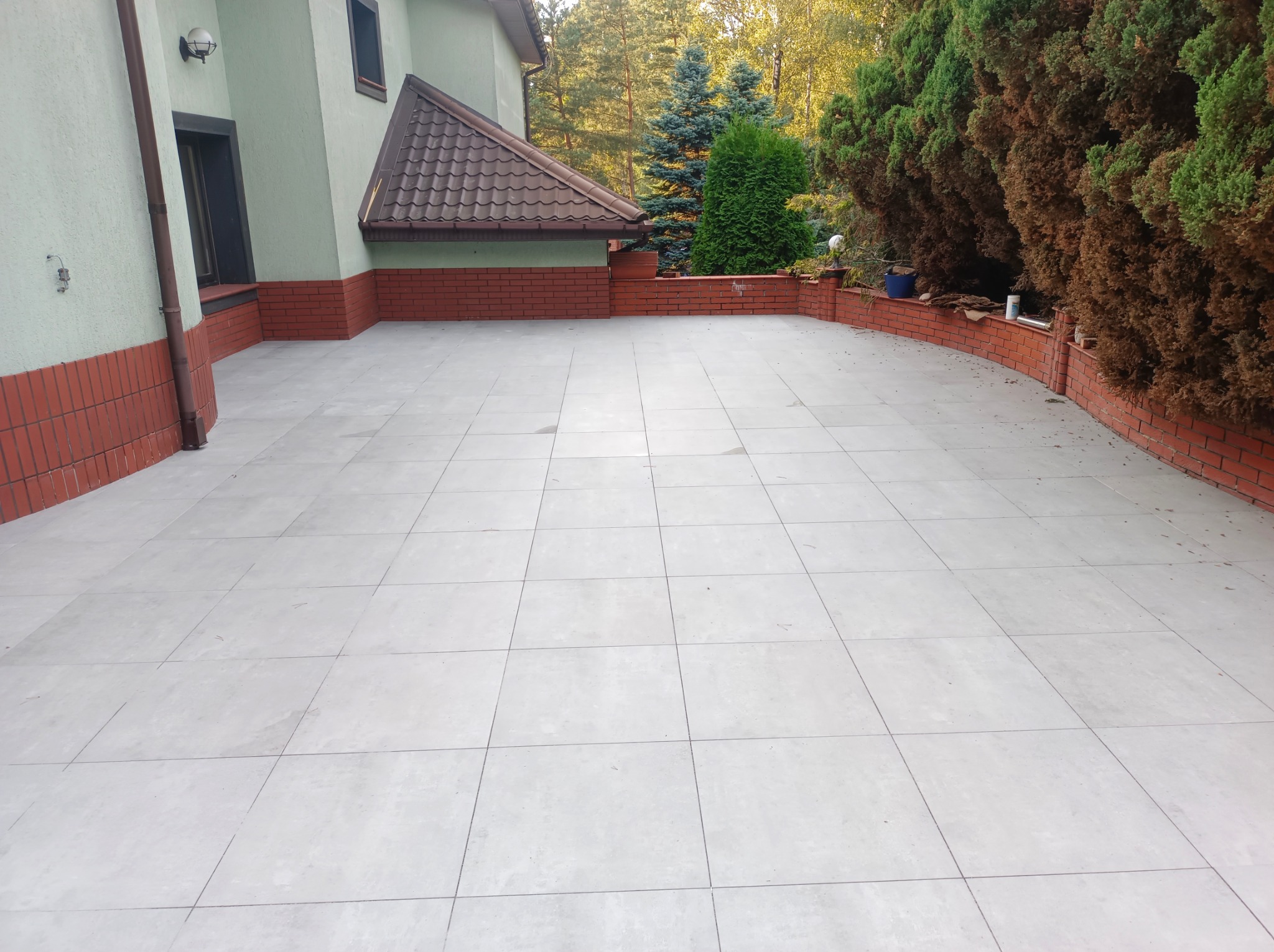 Jasnoszara, kwadratowa nawierzchnia z płyt na zewnątrz budynku z ceglaną podmurówką i zielenią w tle, wykonana w regularnym wzorze.