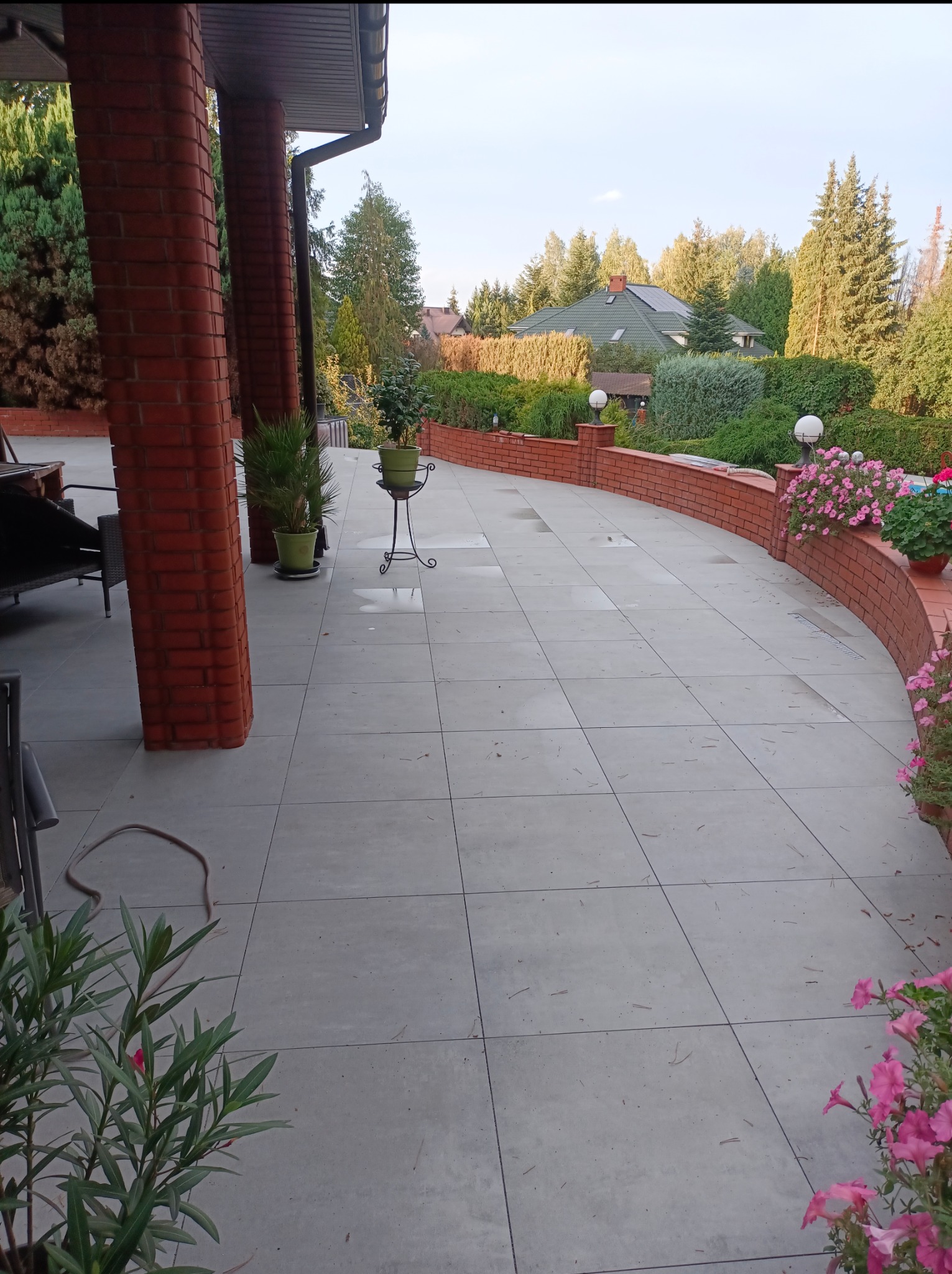 Jasnoszara, kwadratowa nawierzchnia tarasu z roślinami w donicach i ceglanym murem, otoczona zielenią. Widok na dom i ogród w słoneczny dzień.