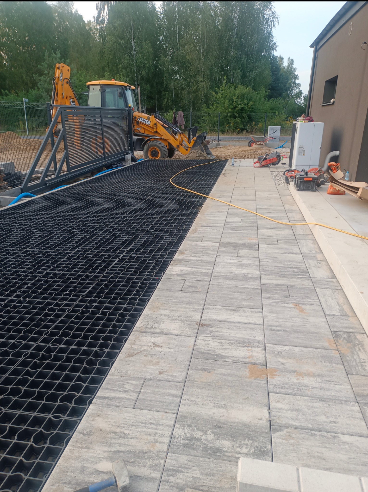 Częściowo ułożona, szara kostka brukowa sąsiaduje z czarną kratką trawnikową. W tle koparka JCB i dom z oknem, sprzęt budowlany. Prace brukarskie w toku.