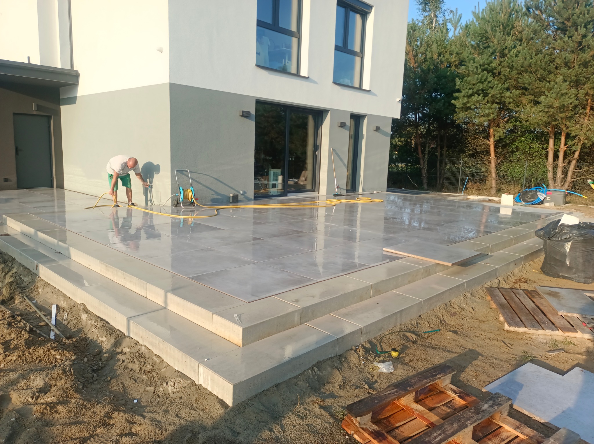 Czyszczenie tarasu z płyt gresowych po montażu, z widocznymi schodami i fragmentem domu w tle. Prace wykończeniowe na zewnątrz budynku.