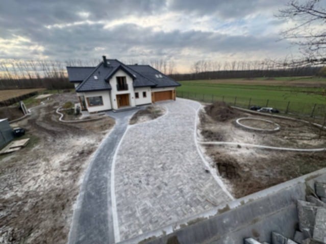 Nowoczesny dom jednorodzinny z podjazdem wyłożonym szarą kostką brukową w jodełkę, obrzeża z jasnej kostki. Widok z góry na posesję w jesiennej scenerii.