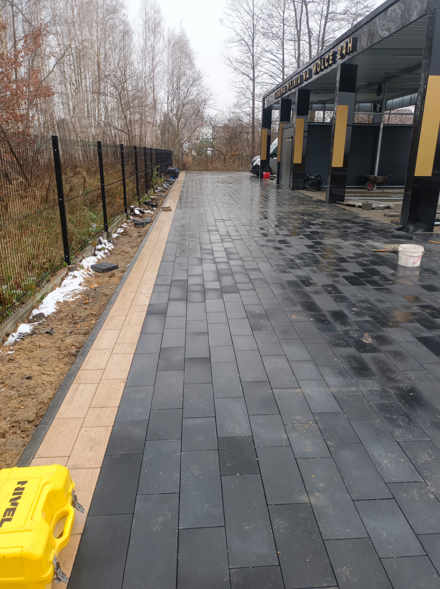 Ułożona, czarna kostka brukowa z jasnym obrzeżem, prowadząca do myjni samochodowej. Widoczny żółty kufer narzędziowy i częściowo stopiony śnieg przy ogrodzeniu.