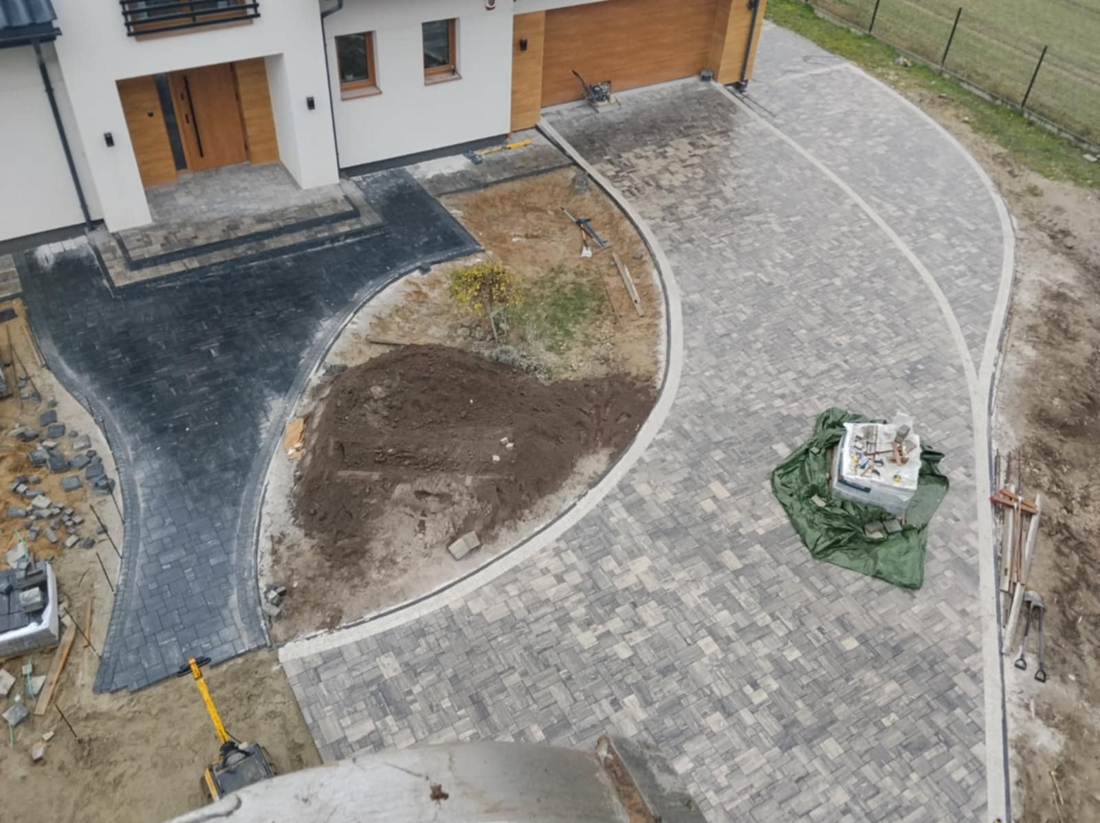 Częściowo ułożona, zakrzywiona nawierzchnia z kostki brukowej w dwóch odcieniach szarości, z widocznym stosem kostki i narzędziami, na tle domu z drewnianymi elementami.