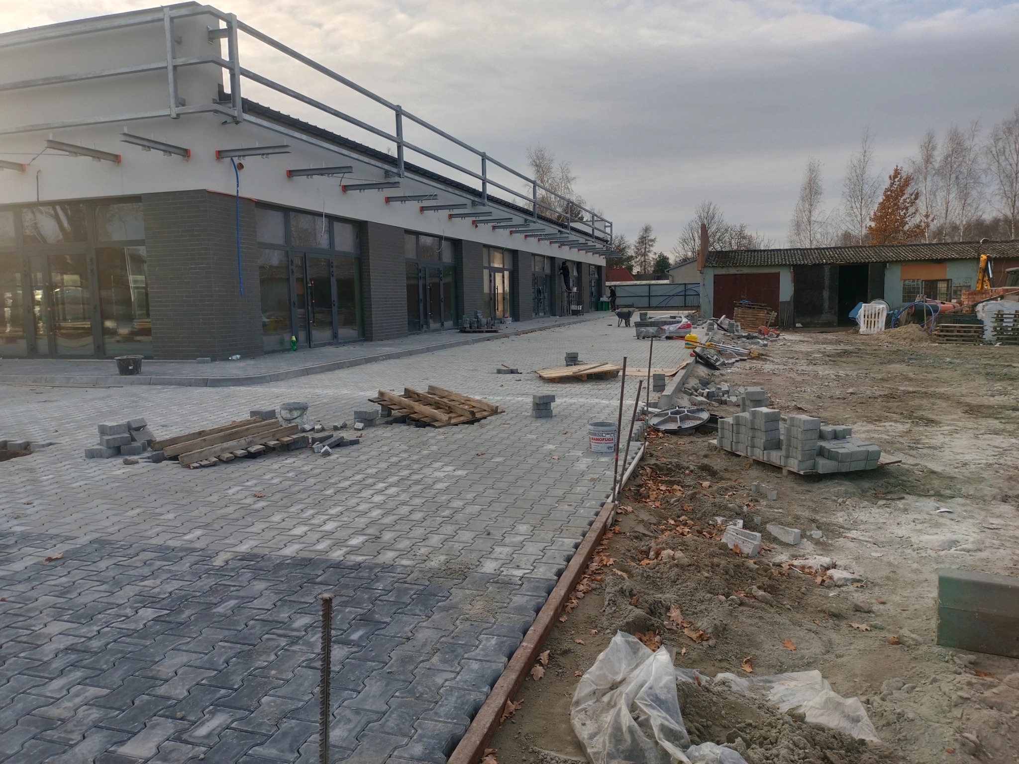 Szara kostka brukowa ułożona na posesji przy budynku. Widoczne materiały budowlane i narzędzia. Częściowo ułożona nawierzchnia, prace w toku, pochmurne niebo.