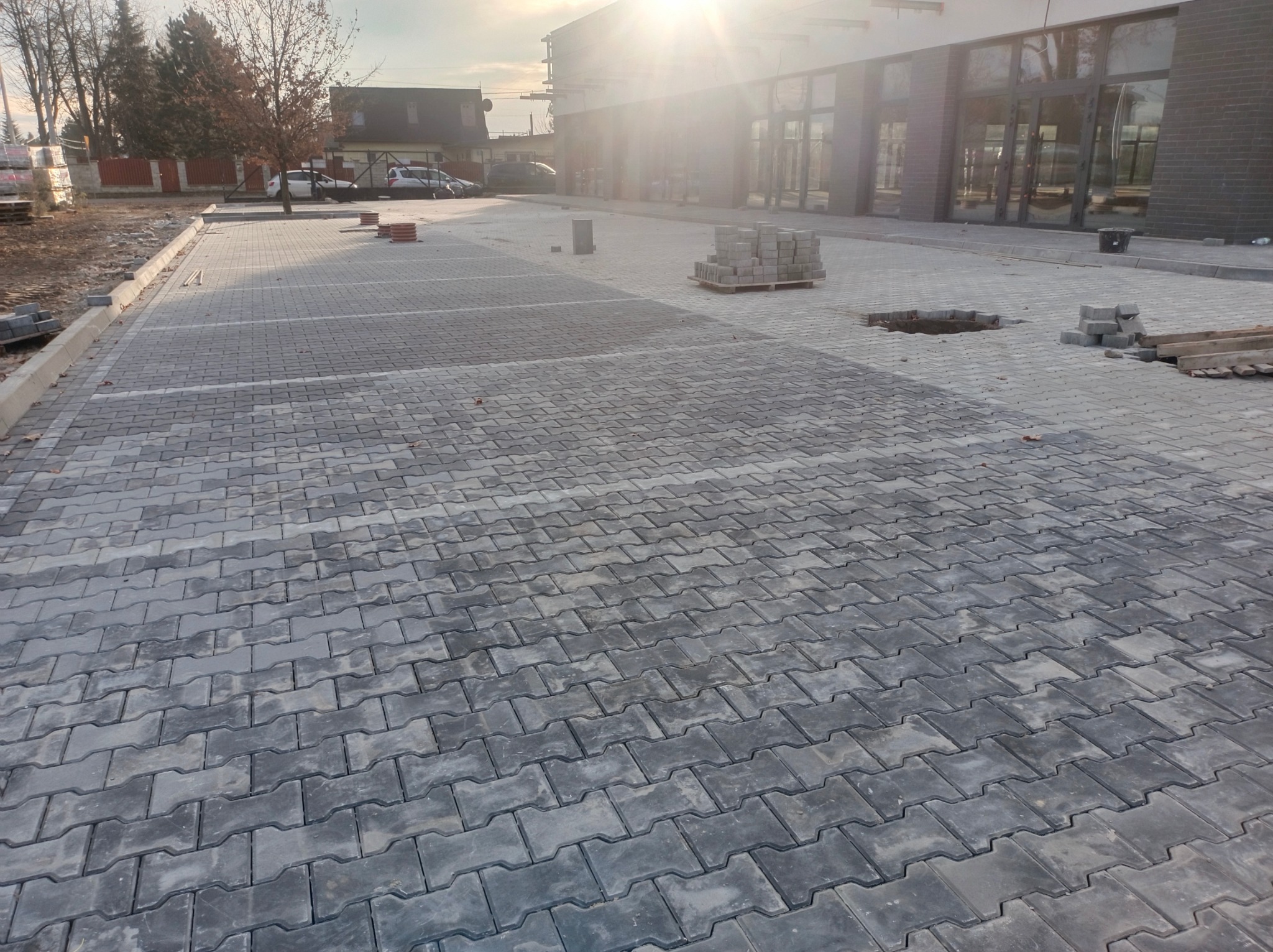 Szara kostka brukowa ułożona na parkingu przed budynkiem w słoneczny dzień. Widoczne palety z kostką i fragmenty niezakończonej nawierzchni.
