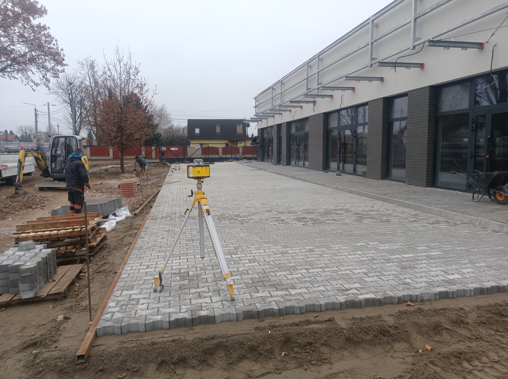 Szara kostka brukowa ułożona przed budynkiem z szarymi oknami. Na pierwszym planie żółty niwelator. W tle pracownicy i mini koparka. Etap prac.