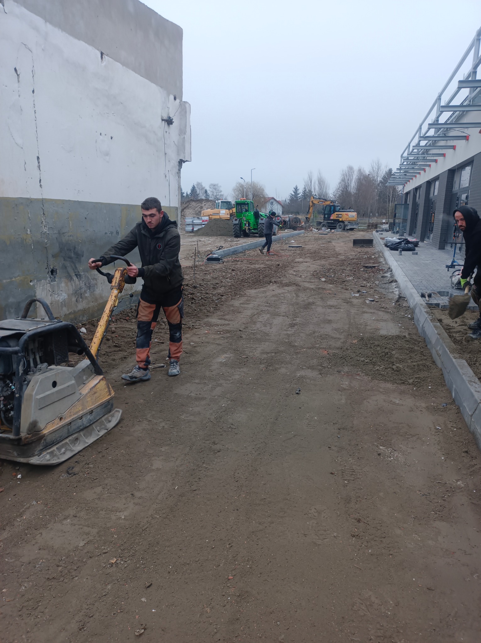 Praca brukarska w toku: ubijanie podłoża pod kostkę brukową zagęszczarką. W tle koparki i przygotowane obrzeża chodnika. Etap prac ziemnych przed ułożeniem kostki.