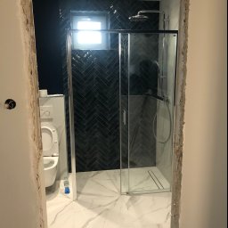 P.N Remont Przemysław Niewiadomski - Łazienka w trakcie remontu: widok przez surową futrynę na kabinę prysznicową z czarnymi płytkami ułożonymi w jodełkę na ścianie i białą marmurową podłogą. Uwagę zwraca butelka wody mineralnej...