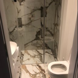 P.N Remont Przemysław Niewiadomski - Wąska łazienka z kabiną prysznicową ze złotą armaturą, umywalką z szafką i podwieszaną toaletą, wykończona płytkami imitującymi marmur Calacatta.