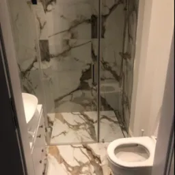 Wąska łazienka z kabiną prysznicową ze złotą armaturą, umywalką z szafką i podwieszaną toaletą, wykończona płytkami imitującymi marmur Calacatta.