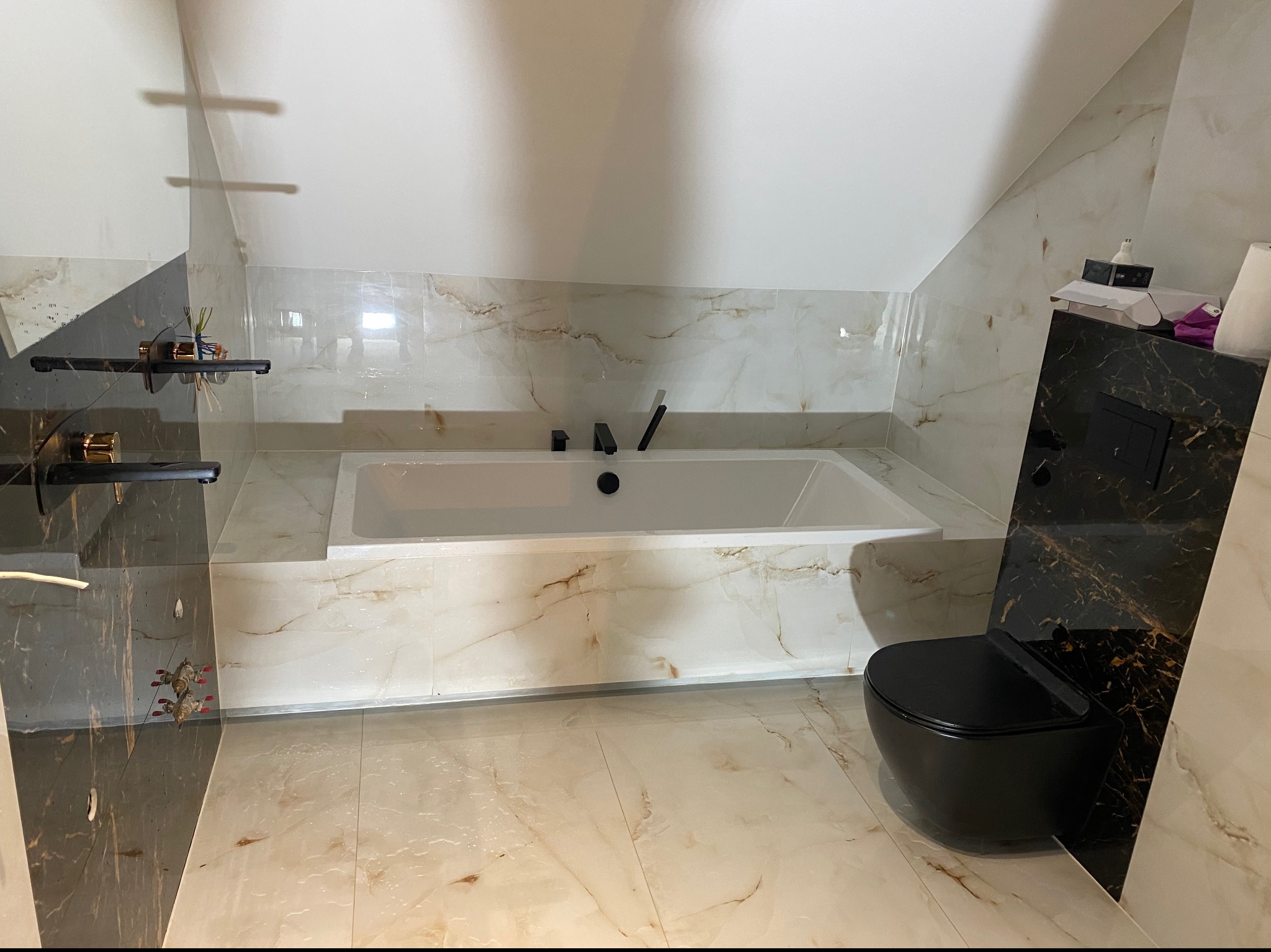 Wykończona łazienka na poddaszu z wanną zabudowaną w płytkach imitujących marmur i wiszącą toaletą w kolorze czarnym, z widocznymi elementami dekoracyjnymi na półce.