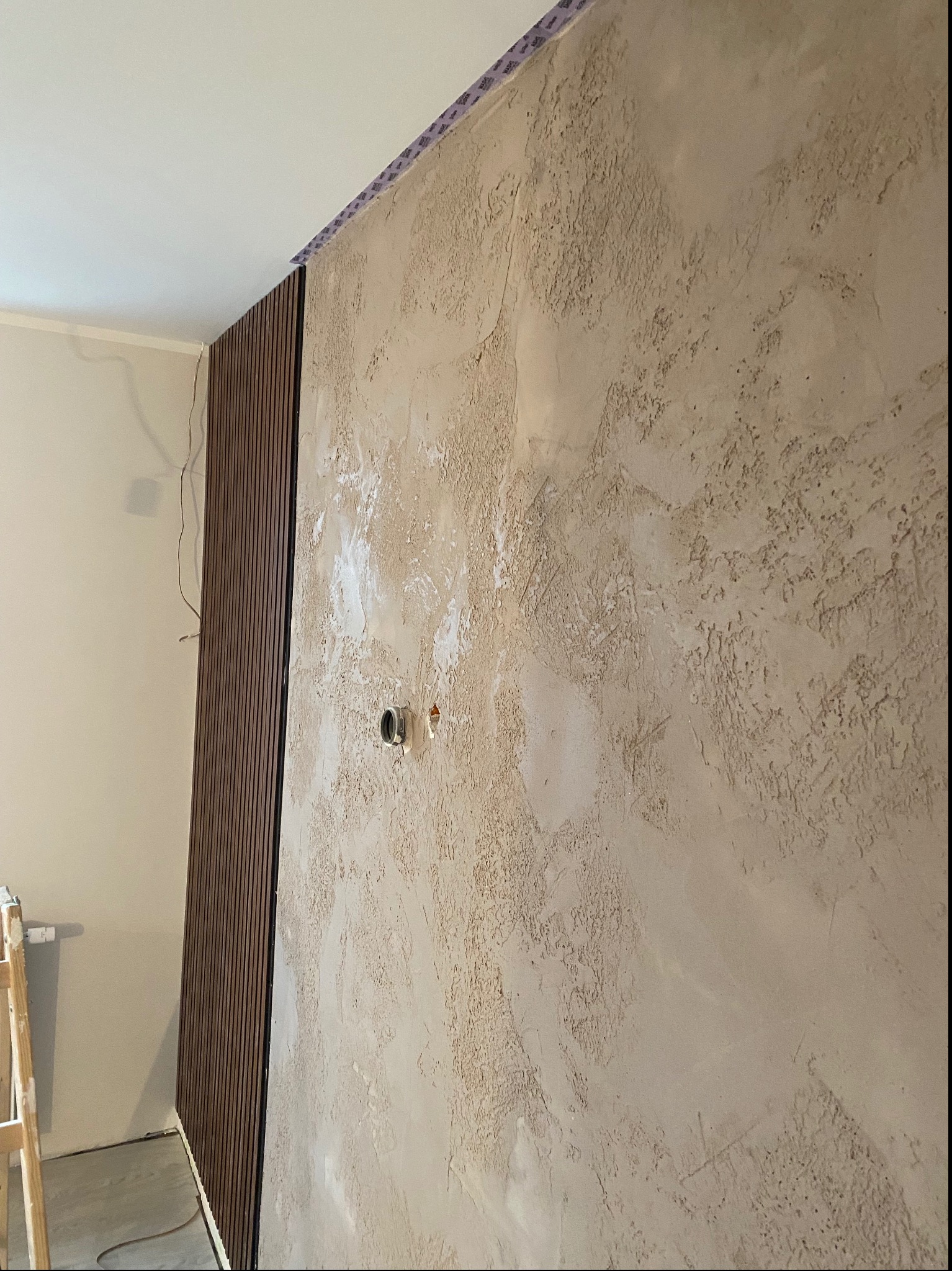 Ściana z dekoracyjną strukturą tynku w odcieniach beżu, obok zamontowane pionowe panele ścienne z ciemnego drewna, widoczne gniazdko elektryczne i fragment drabiny.