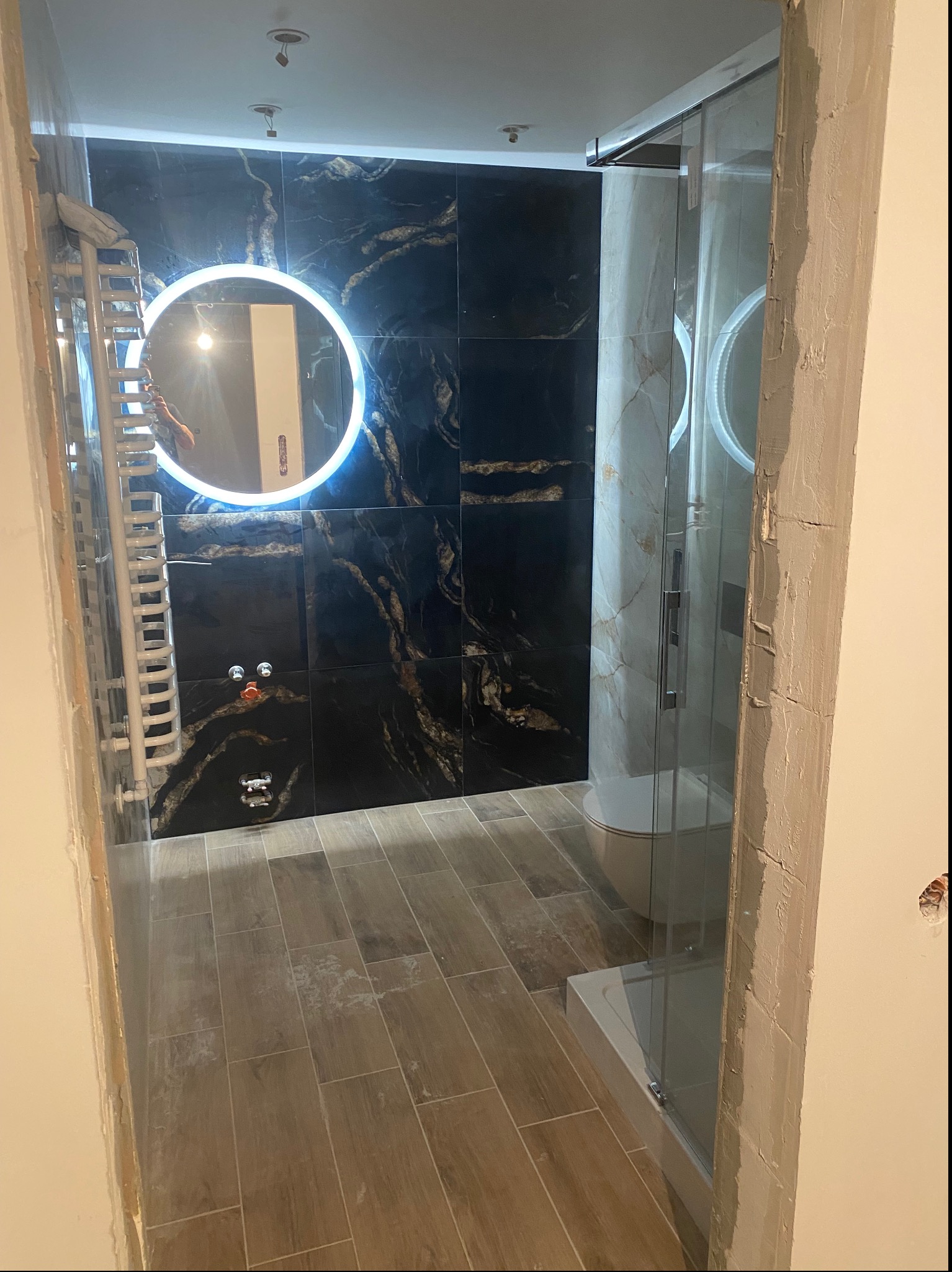 Wnętrze łazienki w trakcie prac wykończeniowych, widoczne czarne płytki imitujące marmur z podświetlanym okrągłym lustrem, kabina prysznicowa oraz grzejnik drabinkowy. Na podłodze położone płytki...