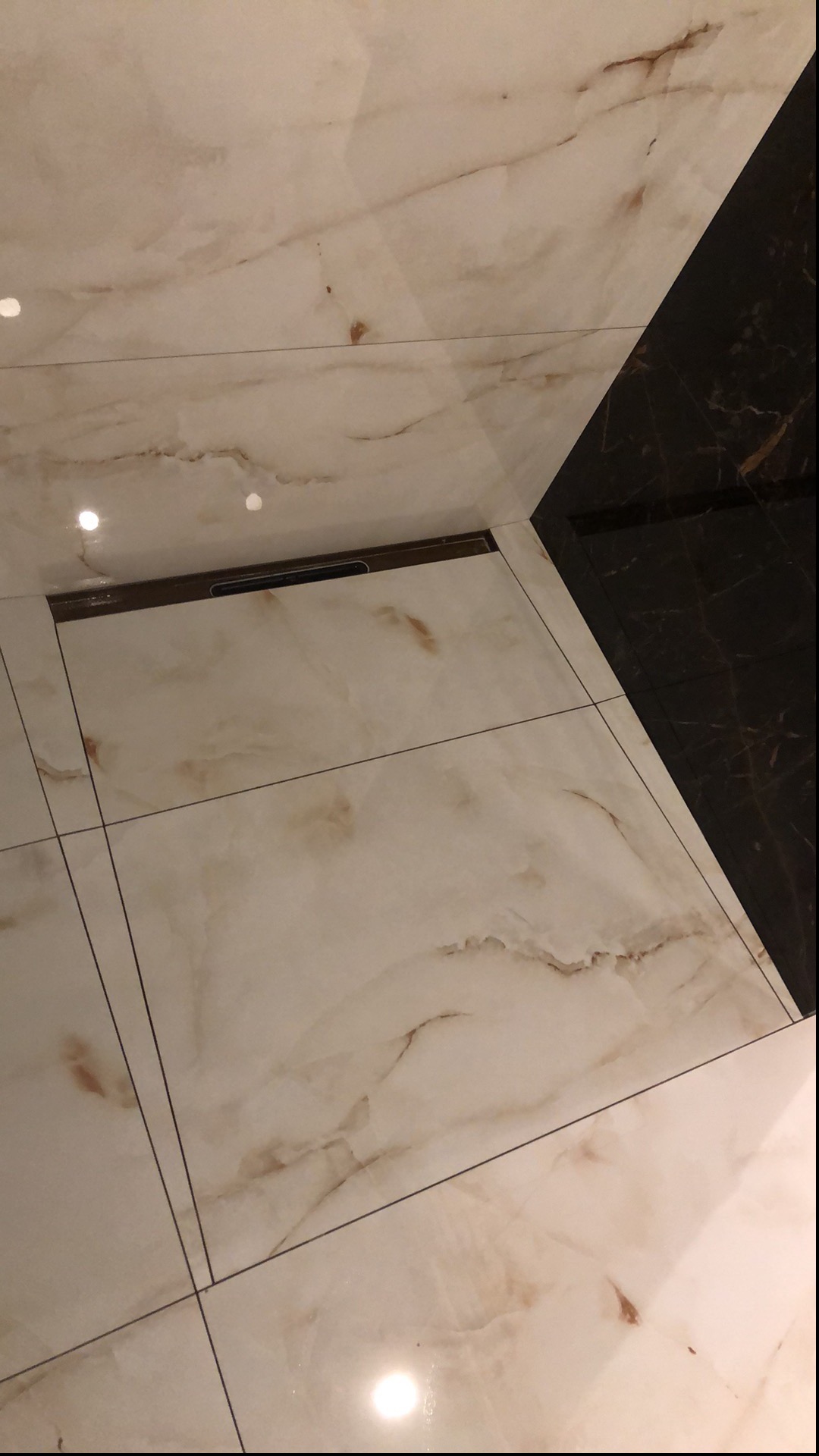 Beżowe i czarne marmurowe płytki podłogowe z odpływem liniowym w łazience, widoczne fugi.